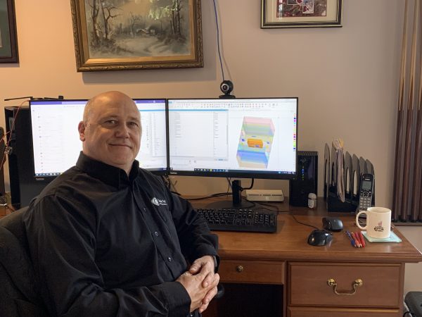 A picture of Jim Korenev smiling in front of his At Home Desk during Covid. There is a Mold Prototype on the screen behind him.