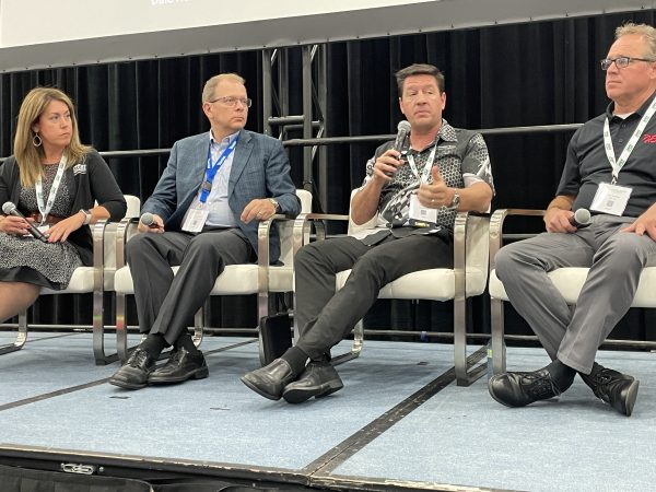 Cavalier Tool's President Brian Bendig is standing on a stage holding a microphone. Brian Bendig participated in a panel at the Injection Molding and Design Expo. He is wearing a Black Cavalier Tool Collared Shirt with checkered flag design on it.