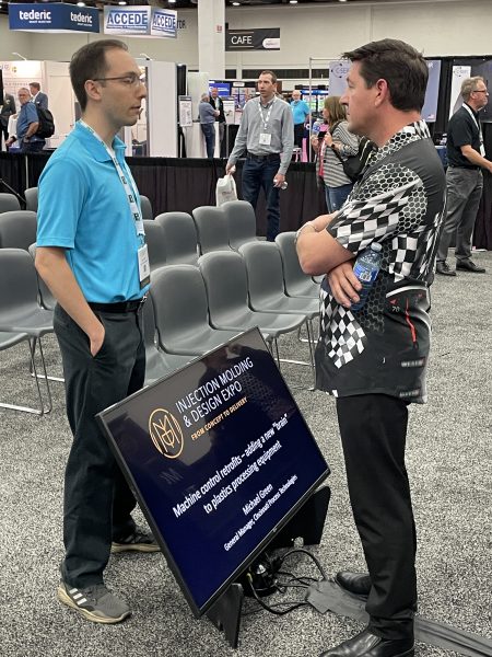 Cavalier Tool President Brian Bendig talking to a member of the audience after participating in a panel at the Injection Molding and Design Expo. Brian is standing with his arms crossed and speaking to a man in a blue shirt.