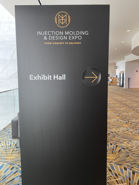 A sign with an arrow pointing to the right. At the top of the sign there is text that says "Injection Molding and Design Expo". Directly underneath that there is smaller orange text that reads "from concept to delivery".