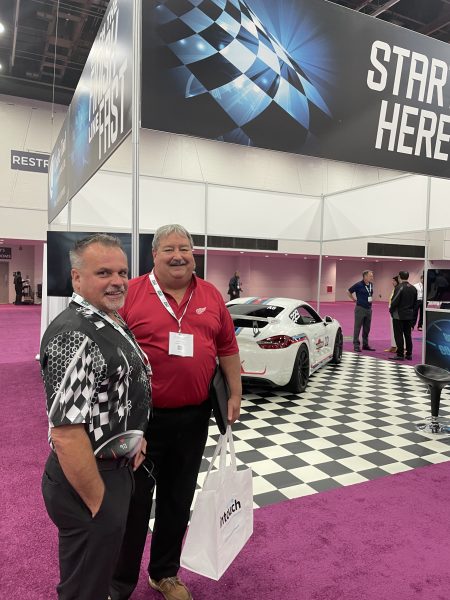 Todd Shepley at the Injection Molding and Design Expo standing in front of Cavalier Tool's booth. Cavalier Tool's President Brian Bendig's White Porsche with a Martini Livery can be seen in the background.