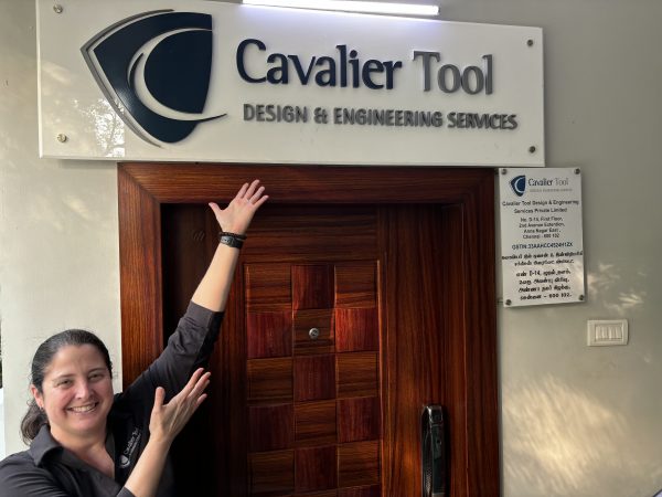 Cavalier CFO Diane Woodiwiss stands in at the front door of one of the Cavalier Tool India locations. She is gesturing towards the Cavalier Tool sign over the door.